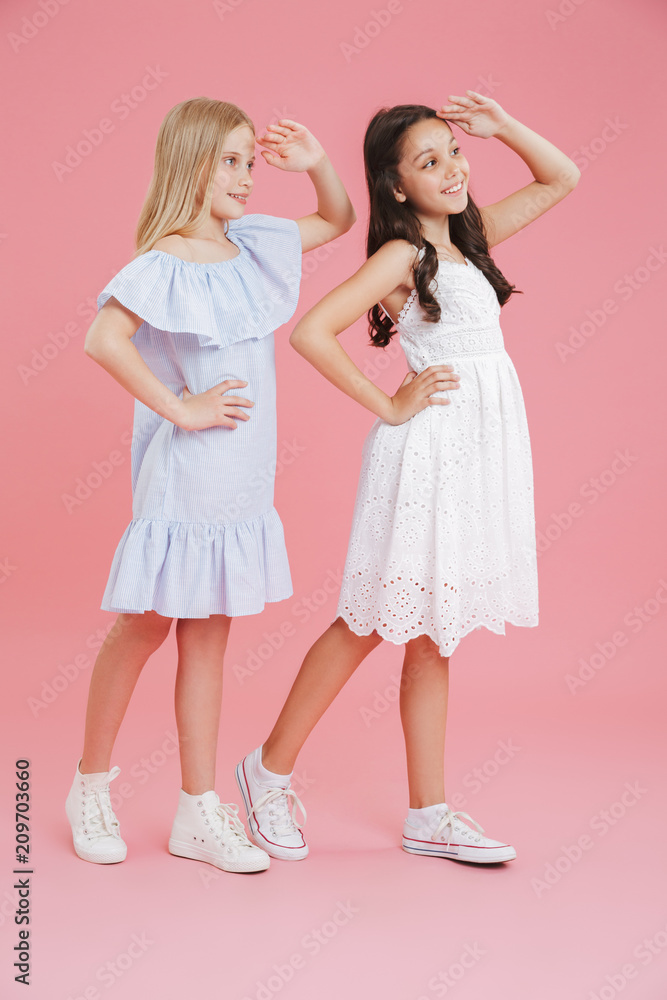 Full length photo of brunette and blonde girls wearing dresses smiling and looking far away with hands at their foreheads, isolated over pink background Full length photo of brunette and blonde girls wearing dresses smiling and looking far away with hands at their foreheads, isolated over pink background