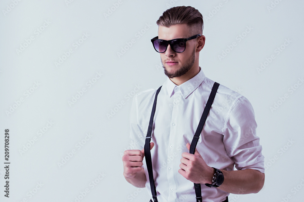Portrait of stylish and elegant man in classical wear and sunglasses  keeping suspenders in hands while standing against gray background. Men's  eyewear fashion and style. Handsome man posing in studio Stock Photo