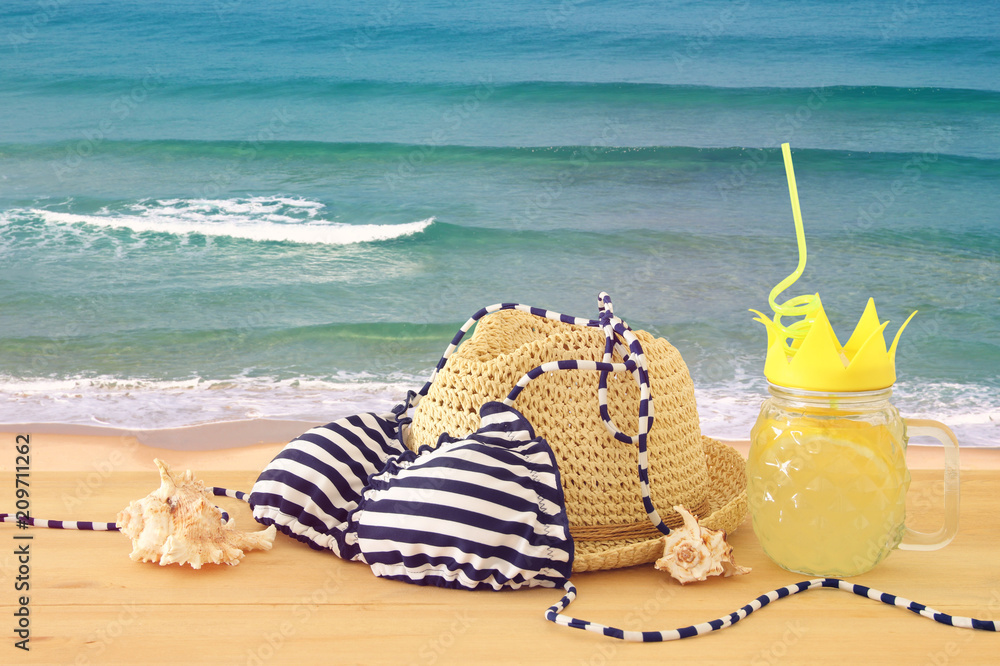 Image of fresh lemonade drink in cute pineapple shape glass with twisted straw next to bikini swimsuit and beach fedora hat over wooden table.