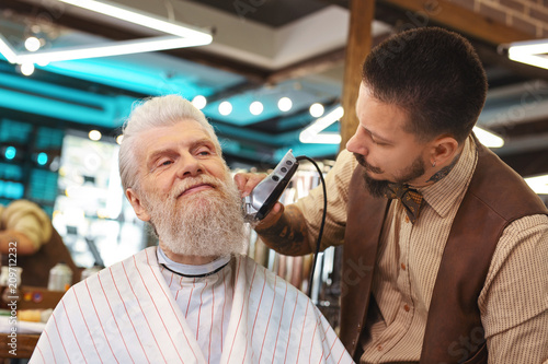 So glad. Professional hairdresser holding hair clipper while modelling modern form of beard