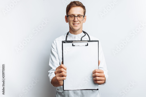 A young doctor shows a folder with a blank sheet on the camera, on a white background, for advertising and inserting text photo