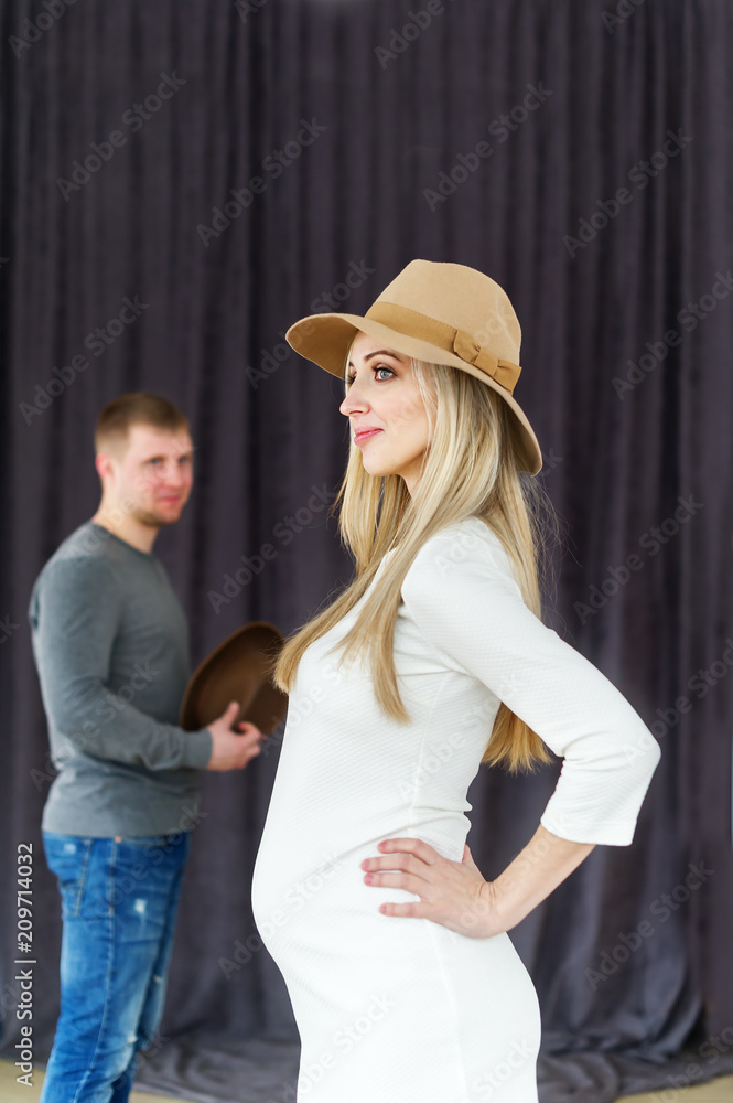 Portrait of the charming pregnant woman in a felt hat and a beautiful white dress. Her husband is at a background Portrait of the charming pregnant woman in a felt hat and a beautiful white dress. Her husband is at a background