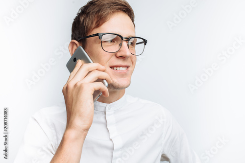 Portrait of young attractive guy in glasses talking on phone and depicting joy, in white shirt isolated on white background, for advertising, text insertion