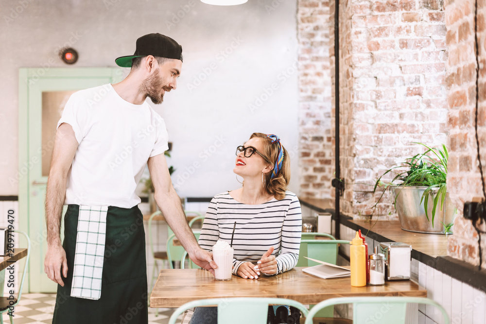 Waiter in black cap giving milkshake to pretty smiling lady in eyeglasses which happily looking at him while sitting at the table in cafe Waiter in black cap giving milkshake to pretty smiling lady in eyeglasses which happily looking at him while sitting at the table in cafe
