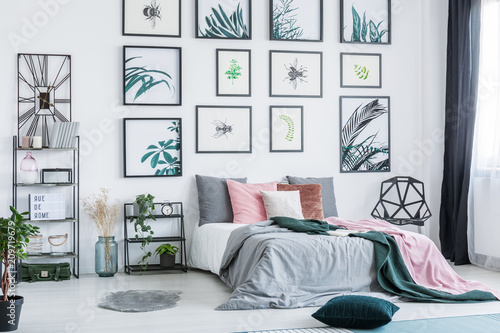 Gallery with simple posters hanging on the wall in bright bedroom interior with many pillows on bed, fresh plants and plastic chair standing next to window with drapes
