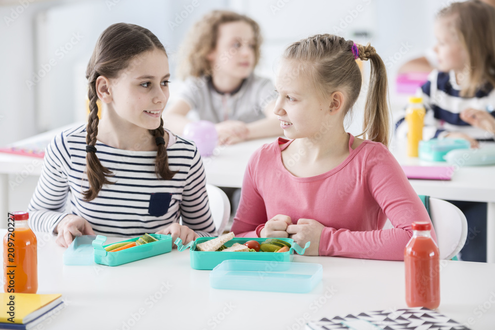 Front view of two girls sitting by a school desk with opened lunch boxes with healthy vegetables and sandwiches. Bottles of orange and tomato juice on a desk. Other children in blurred background Front view of two girls sitting by a school desk with opened lunch boxes with healthy vegetables and sandwiches. Bottles of orange and tomato juice on a desk. Other children in blurred background