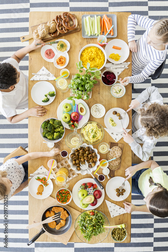 Top view of children eating healthy homemade meal. Wooden table with dishes full of colorful cooked food, eco vegetables, fruit, wholegrain bread rolls and pretzels.