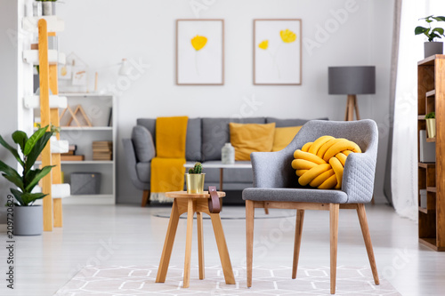 Yellow knot cushion on a gray armchair standing next to a wooden stool with a cactus on it in cozy living room interior. Real photo with blurred background