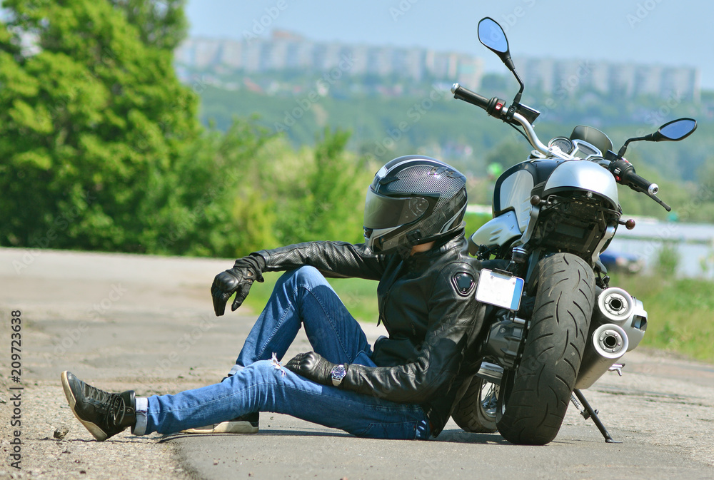 Biker in outfit sits next to his motorcycle on the ground. Motorcyclist  rests near the motorbike. Stock Photo | Adobe Stock