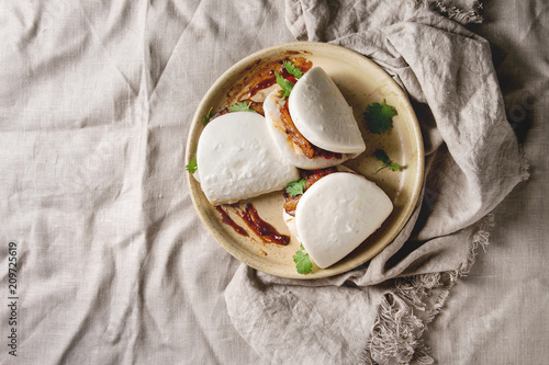 Asian sandwich steamed gua bao buns with pork belly, greens and vegetables served in ceramic plate over linen tablecloth. Asian style fast food dinner. Flat lay, space photo