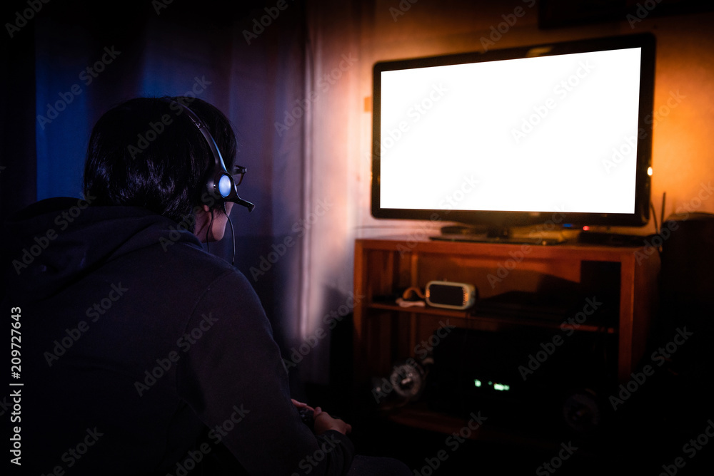 Young male gamer with glasses and headset playing video game at home in the dark room using game console controller watching at LED TV. Gaming and entertainment concepts Young male gamer with glasses and headset playing video game at home in the dark room using game console controller watching at LED TV. Gaming and entertainment concepts