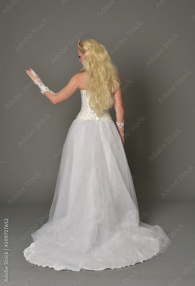 Fototapeta premium full length portrait of blonde girl wearing white gown, standing pose with back to the camera. grey studio background.