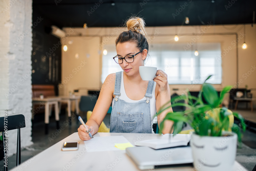 Student girl studying in the cafe. Writing with pen and holding a cup. Student girl studying in the cafe. Writing with pen and holding a cup.