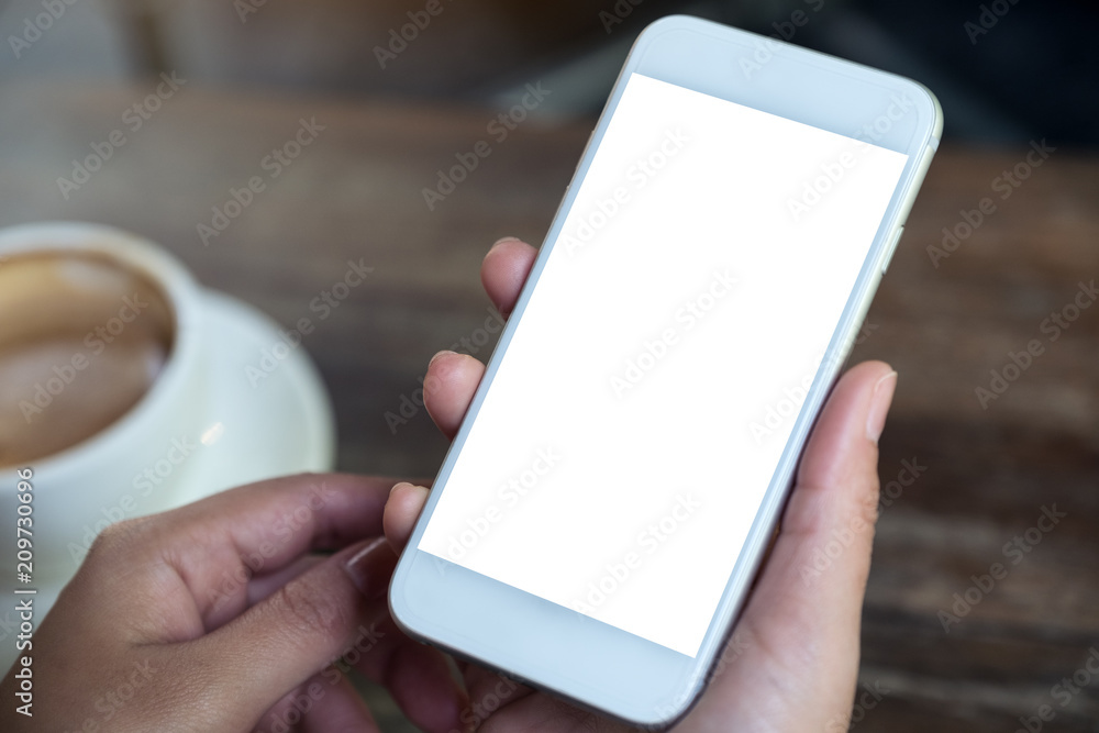 Mockup image of hands holding white mobile phone with blank desktop screen with coffee cup on wooden table in cafe