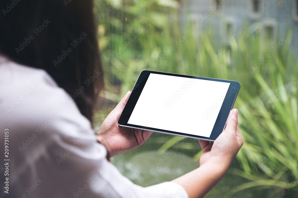 Mockup image of woman's hands holding black tablet pc with blank white desktop screen and green nature background