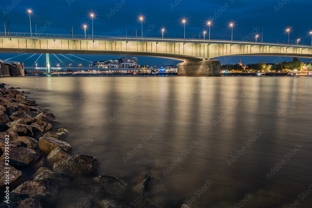 Deutzer Brücke während der blauen Stunde bei Nacht mit den Kranhäusern und der Severinsbrücke im Hintergrund, im Vordergrund Steine
