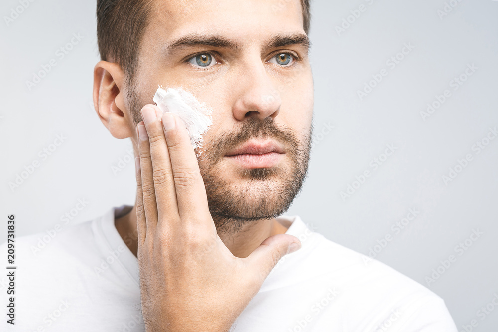 Skin care. Handsome young shirtless man applying cream at his face and looking at himself with smile while standing over gray background and looking at camera. Close-Up. Space for text. Skin care. Handsome young shirtless man applying cream at his face and looking at himself with smile while standing over gray background and looking at camera. Close-Up. Space for text.
