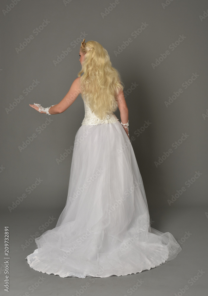 Naklejka premium full length portrait of blonde girl wearing white gown, standing pose with back to the camera. grey studio background.