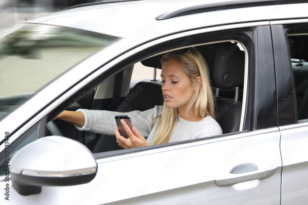 Hübsche junge blonde Frau fährt mit einem Auto und schaut dabei auf ihr Handy