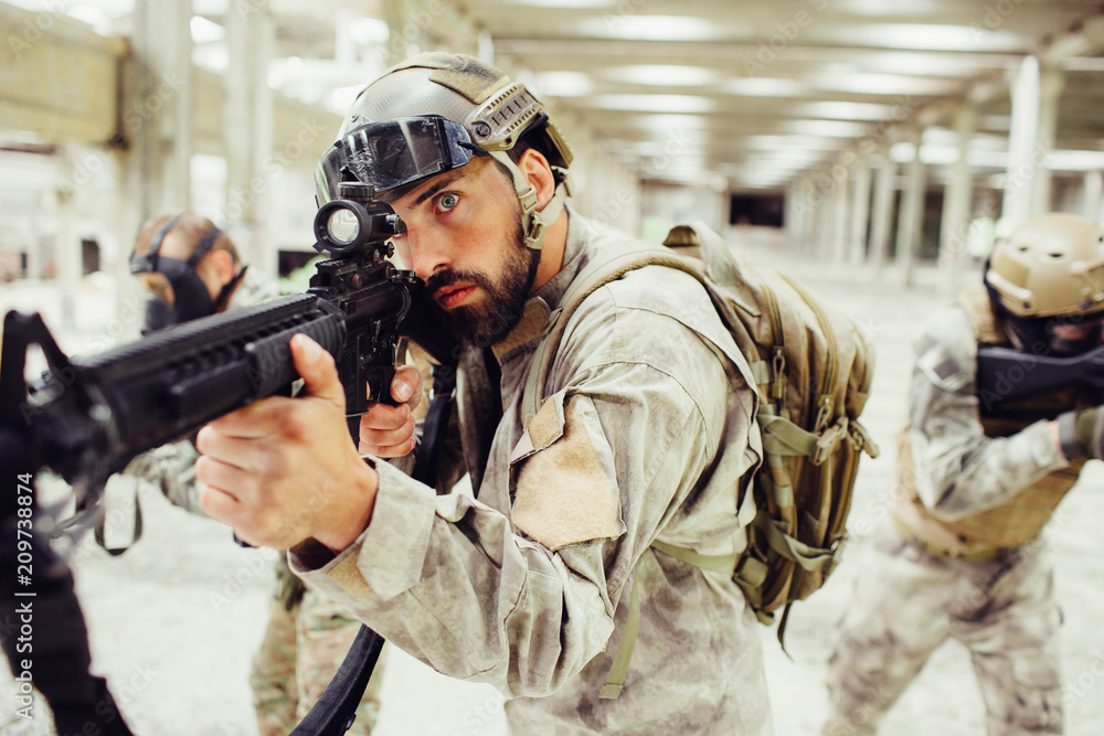 Brave and serious soldier is standing with his combads in a bright and shiny long room. Bearded guy is taking aim while the other two warriors are looking to different sides. They are working as a Brave and serious soldier is standing with his combads in a bright and shiny long room. Bearded guy is taking aim while the other two warriors are looking to different sides. They are working as a