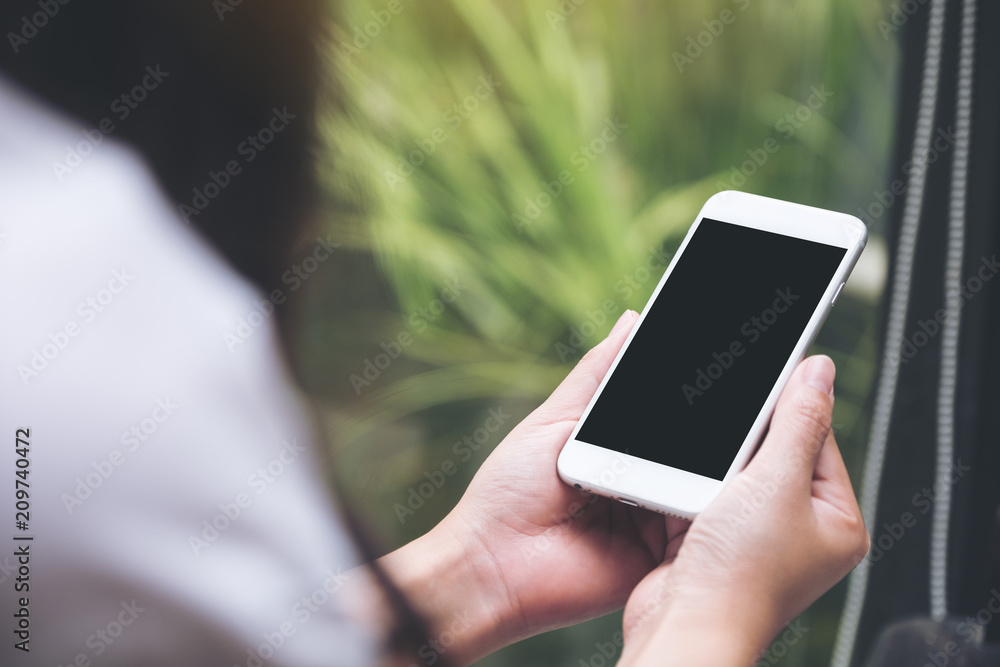 Mockup image of a woman holding white smart phone with blank black desktop screen with blur green nature background