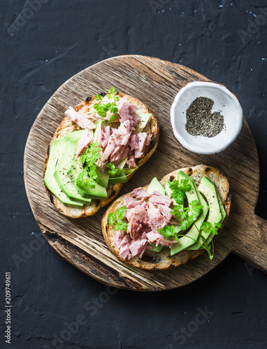 Sandwiches with cream cheese, avocado and tuna fish on wooden cutting board on dark background, top view. Healthy breakfast or snack