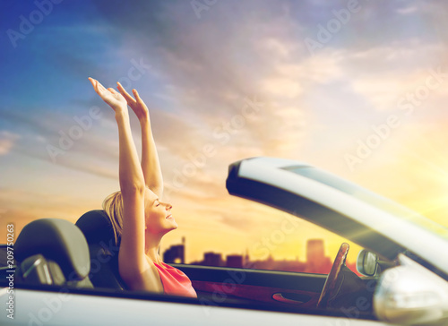 travel, summer holidays, road trip and people concept - happy young woman in convertible car enjoying sunset over city background