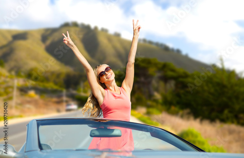 travel, summer holidays, road trip and people concept - happy young woman wearing sunglasses in convertible car showing peace hand sign over big sur hills and road background in california