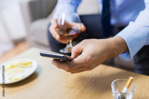 alcoholism, alcohol addiction and technology concept - close up of male hands with smartphone and brandy at home