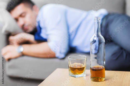 alcoholism, alcohol addiction and people concept - bottle with glass of whiskey on table and sleeping drunk man