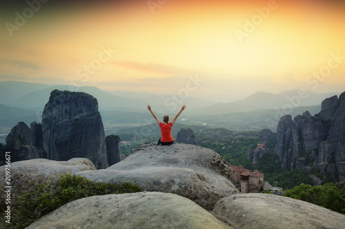 Sunset over Meteora monasteries in Greece. Girl tourist sitting on top of the rock and enjoying beautiful panoramic landscape of Meteora area