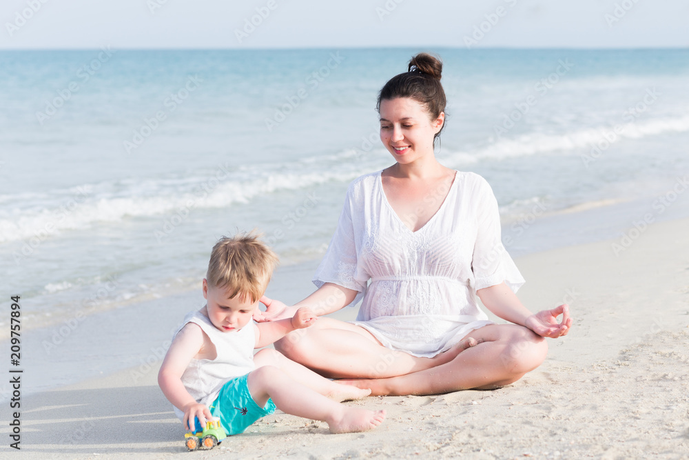 Young pregnant mother in white dress and her little toddler son playing and relaxing on the sea beach with sand at summer vacation time. Pregnancy, happy family, parenthood concept Young pregnant mother in white dress and her little toddler son playing and relaxing on the sea beach with sand at summer vacation time. Pregnancy, happy family, parenthood concept