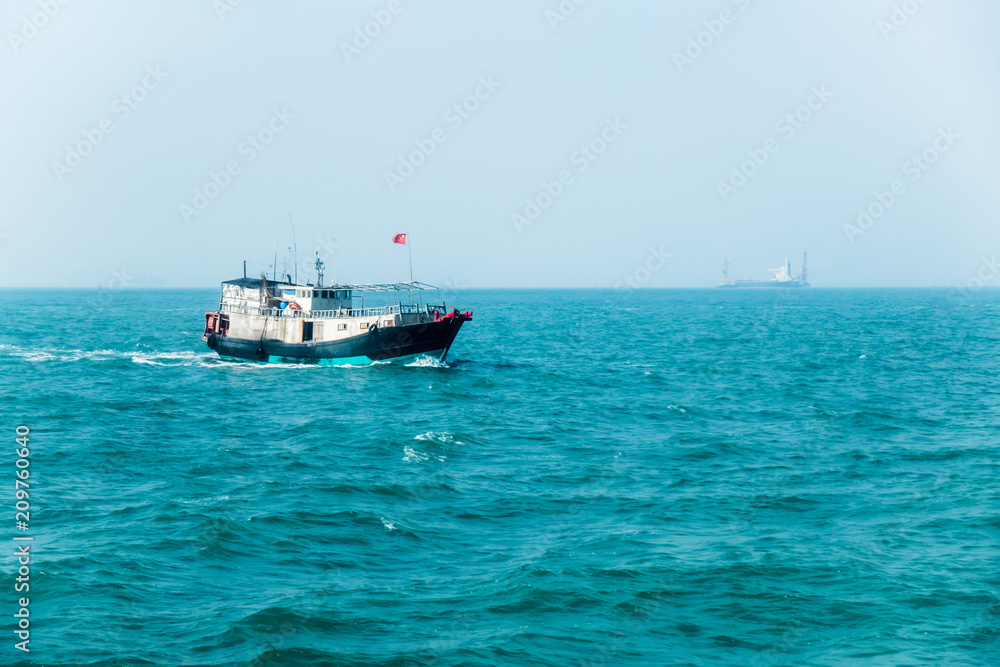 Small old fishing boat with Chinese flag in South China Sea near Hong Kong. Large container ship with cranes in haze on horizon line. Sunny summer day. Fishing industry. 