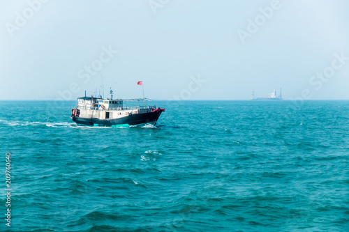 Small old fishing boat with Chinese flag in South China Sea near Hong Kong. Large container ship with cranes in haze on horizon line. Sunny summer day. Fishing industry. 