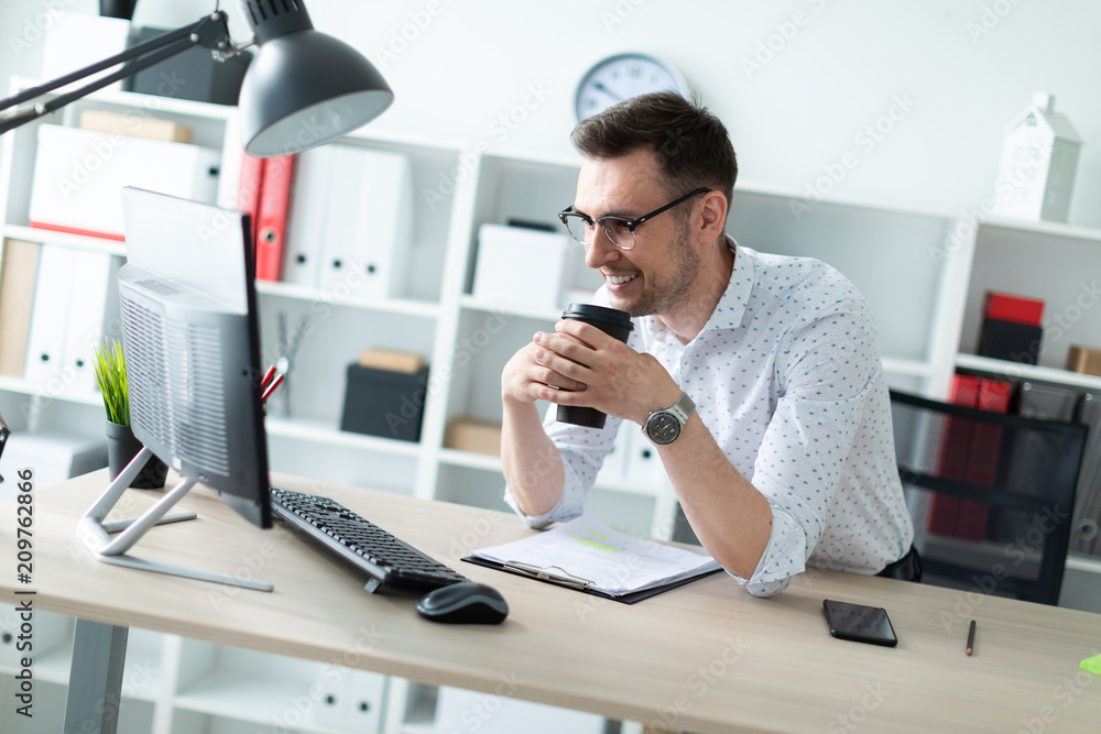 A young man in glasses stands near a table in the office holding a glass of coffee. A young man in glasses stands near a table in the office holding a glass of coffee.