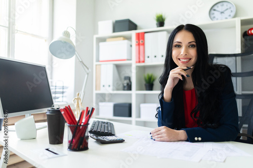 A beautiful young girl sits in the office at the table and holds a pencil in her hand.