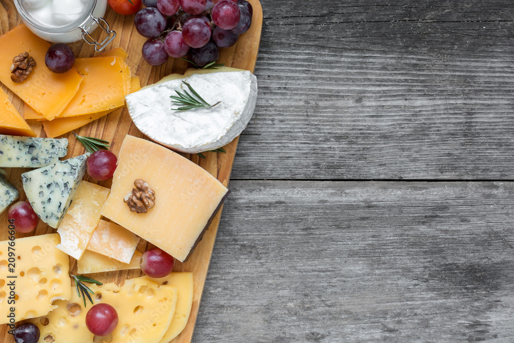 Assorted cheeses on wooden board plate served with walnuts, grapes and rosemary on rustic wood background, top view