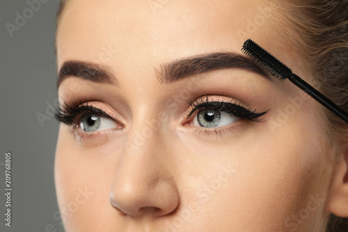 Young woman correcting shape of eyebrows on grey background, closeup