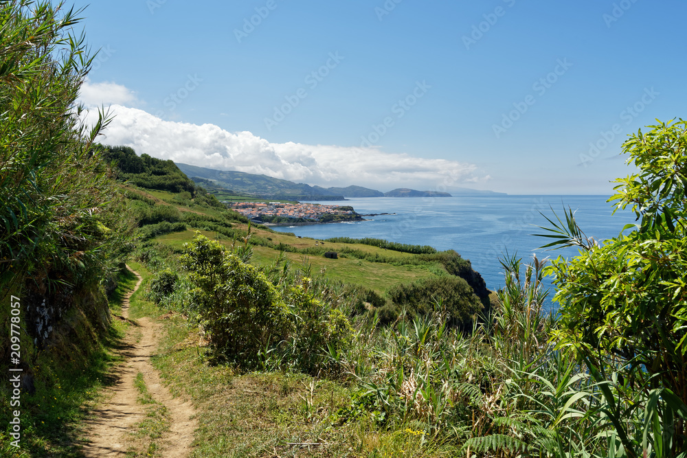 Coastal walk on the Azores island of Sao Miguel in sunny weather, the view follows the trail past pastures to a village and mountains with clouds - Location: Azores, island of Sao Miguel Coastal walk on the Azores island of Sao Miguel in sunny weather, the view follows the trail past pastures to a village and mountains with clouds - Location: Azores, island of Sao Miguel