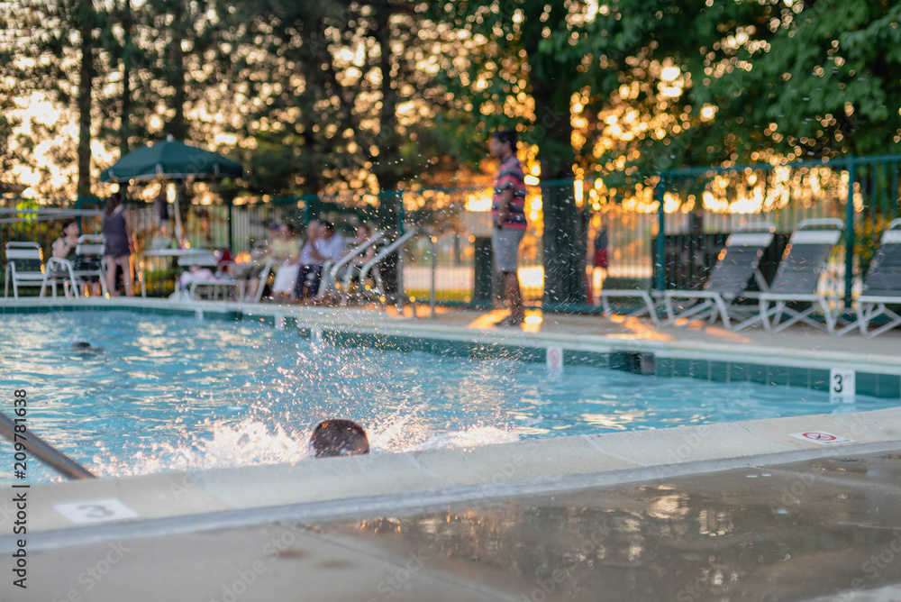 Little boy splashes in swimming pool while adults sit in lawn chairs