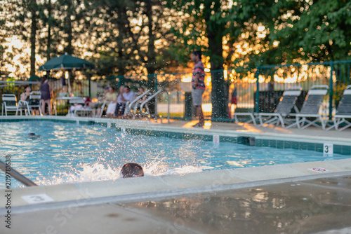 Little boy splashes in swimming pool while adults sit in lawn chairs