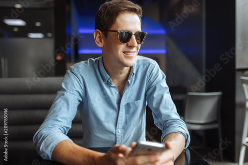 Happy young man in glasses, with phone in hand, sitting in cafe, suitable for advertising, text insertion