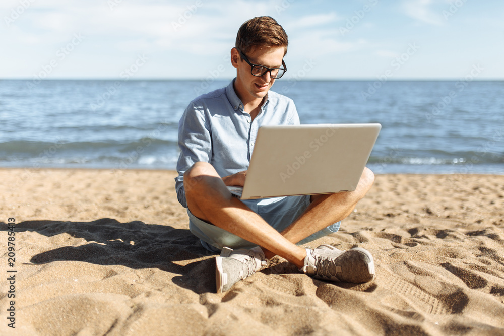 Young guy with glasses, working on his laptop on the beach, work on vacation, suitable for advertising, text insertion
