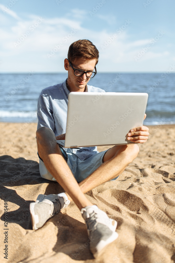 Young guy with glasses, working on his laptop on the beach, work on vacation, suitable for advertising, text insertion