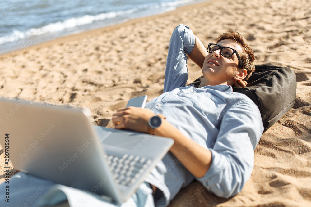 Young guy with glasses, lying on the sand, working on his laptop on the beach, against the sea, working on vacation, suitable for advertising, text insertion