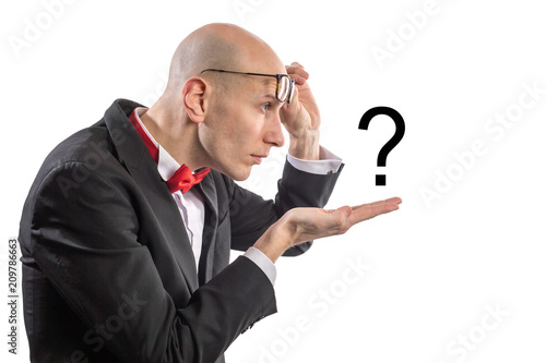 Slim bald elegant nerd with glasses and bow tie looking at question mark on his hand with emotion of bewilderment. Isolated on white.