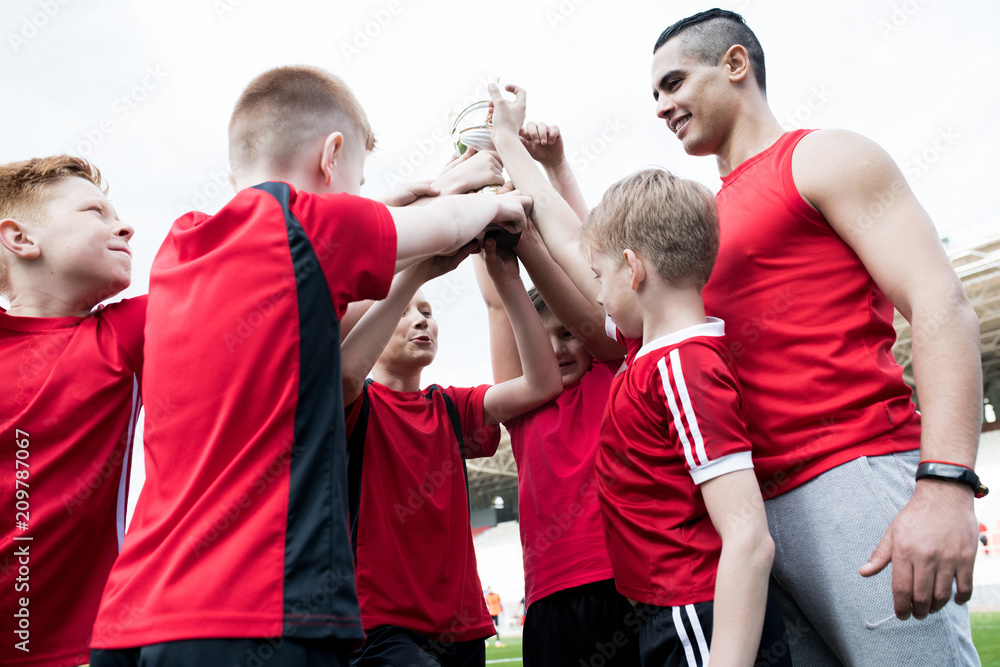 Fototapeta Portrait of happy junior football team holding trophy together and cheering after winning match in outdoor stadium