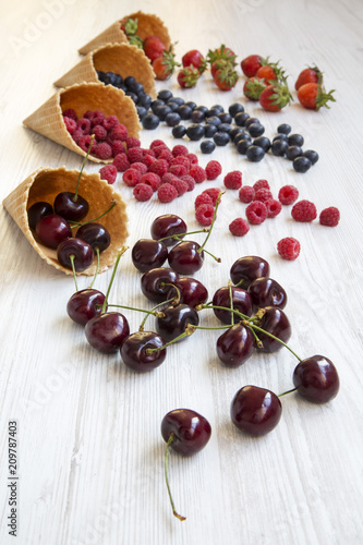 Waffle sweet ice cream cones with raspberries, cherries, strawberries and blueberries over white wooden background, side view.