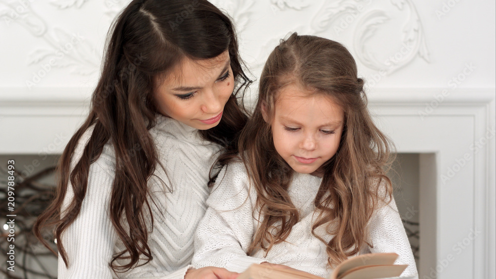 Happy mother and her beautiful daughter in white sweaters reading a book on Christmas eve Happy mother and her beautiful daughter in white sweaters reading a book on Christmas eve