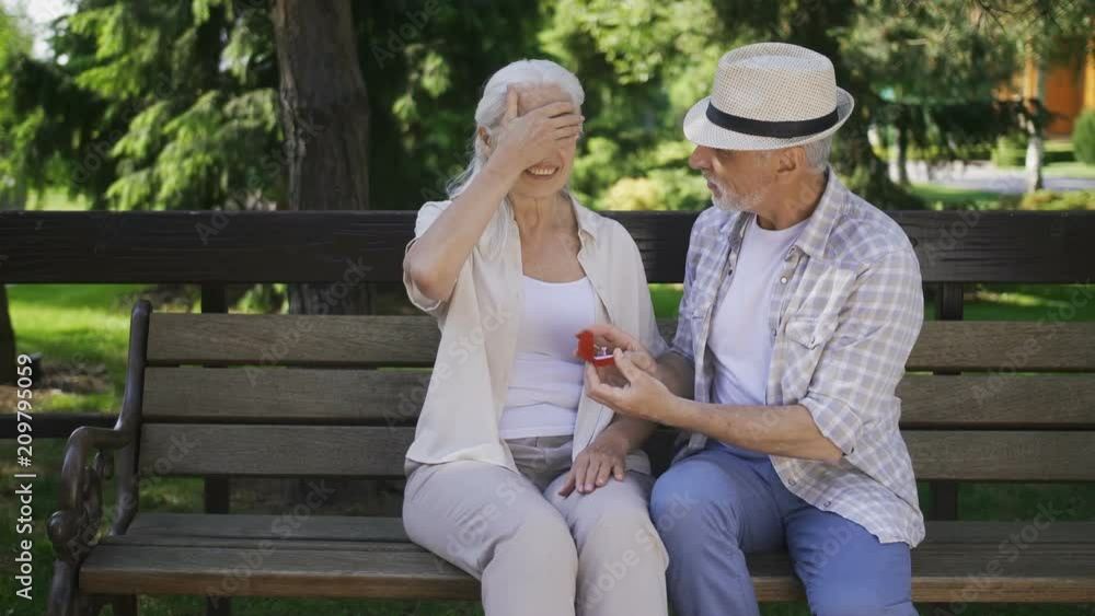 Handsome senior man with gray beard surprising his mature beautiful woman with white hair with marriage proposal while two sitting on a bench. Female with closed eyes getting engagement ring from man Handsome senior man with gray beard surprising his mature beautiful woman with white hair with marriage proposal while two sitting on a bench. Female with closed eyes getting engagement ring from man